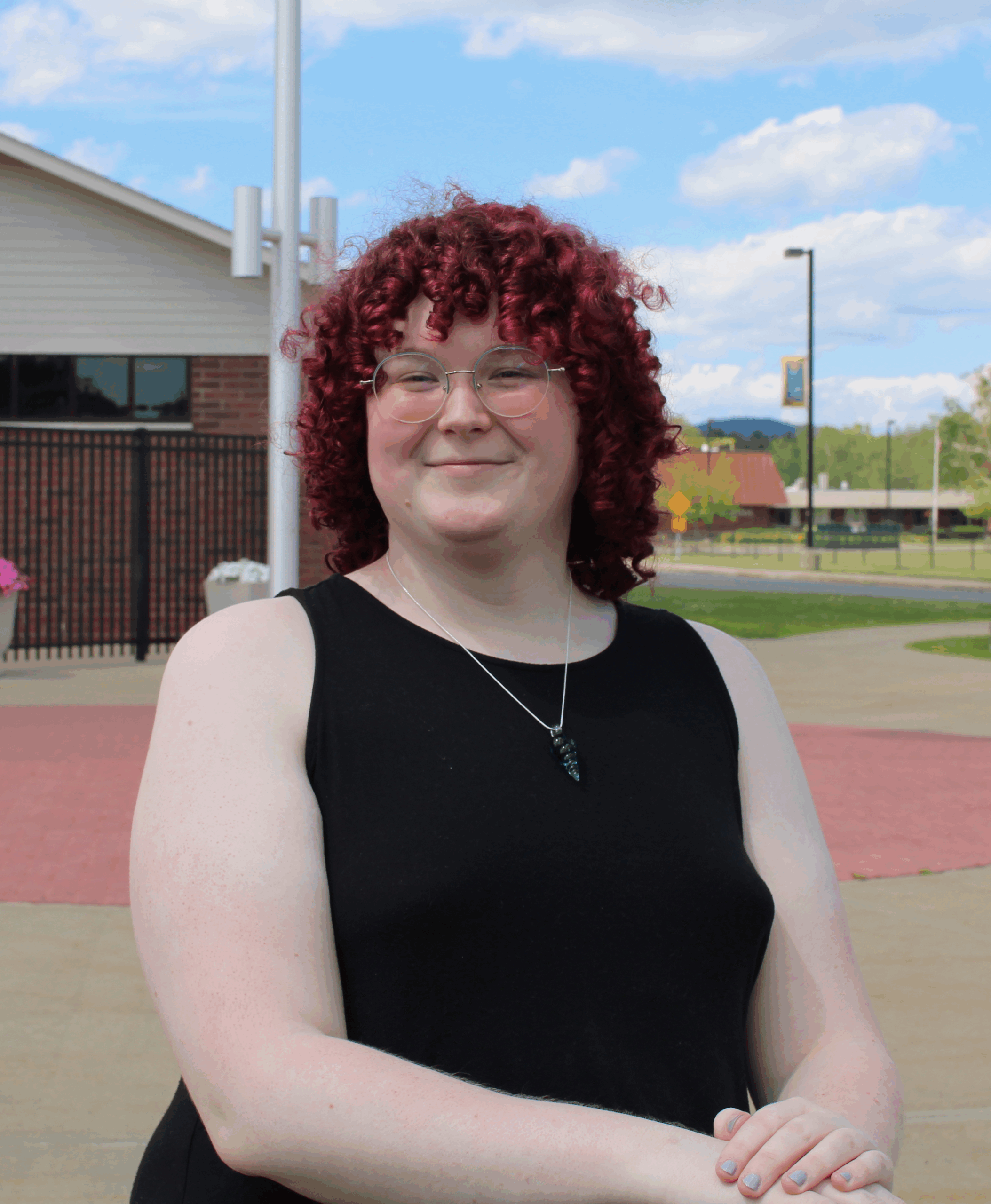 Portrait of a woman named Gwen with red curly hair and she is wearing a black sleeveless dress and a necklace and glasses.